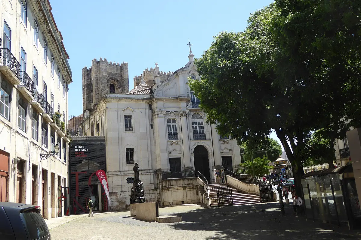 A cobbled square in Lisbon with a cream-coloured baroque church and the crenellated towers of the cathedral rising behind it