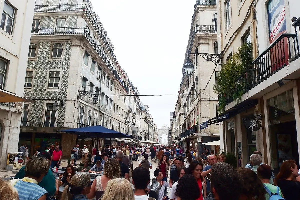 A busy pedestrian street in central Lisbon lined with tall pale buildings, with a triumphal arch visible at the far end